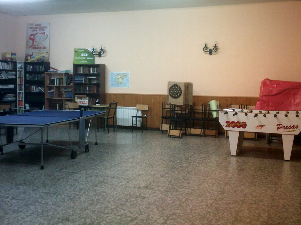 Sala amplia con un futbolín, una diana para dardosm una mesa de pimpón y varias estanterias con libros al fondo a la izquierda.