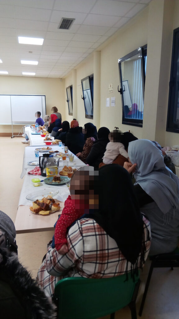 mujeres sentadas ante una larga mesa llena de alimentos, en una sala con una pantalla al fondo