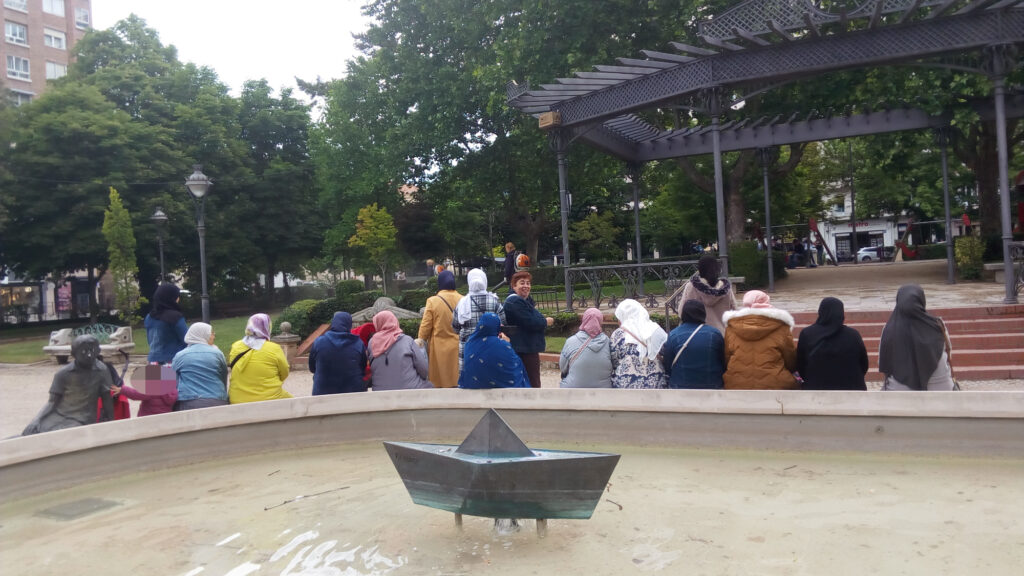 Grupo de mujeres de espaldas en el Poniente, sentadas en la fuente que tiene una escultura de un barco de papel en el centro
